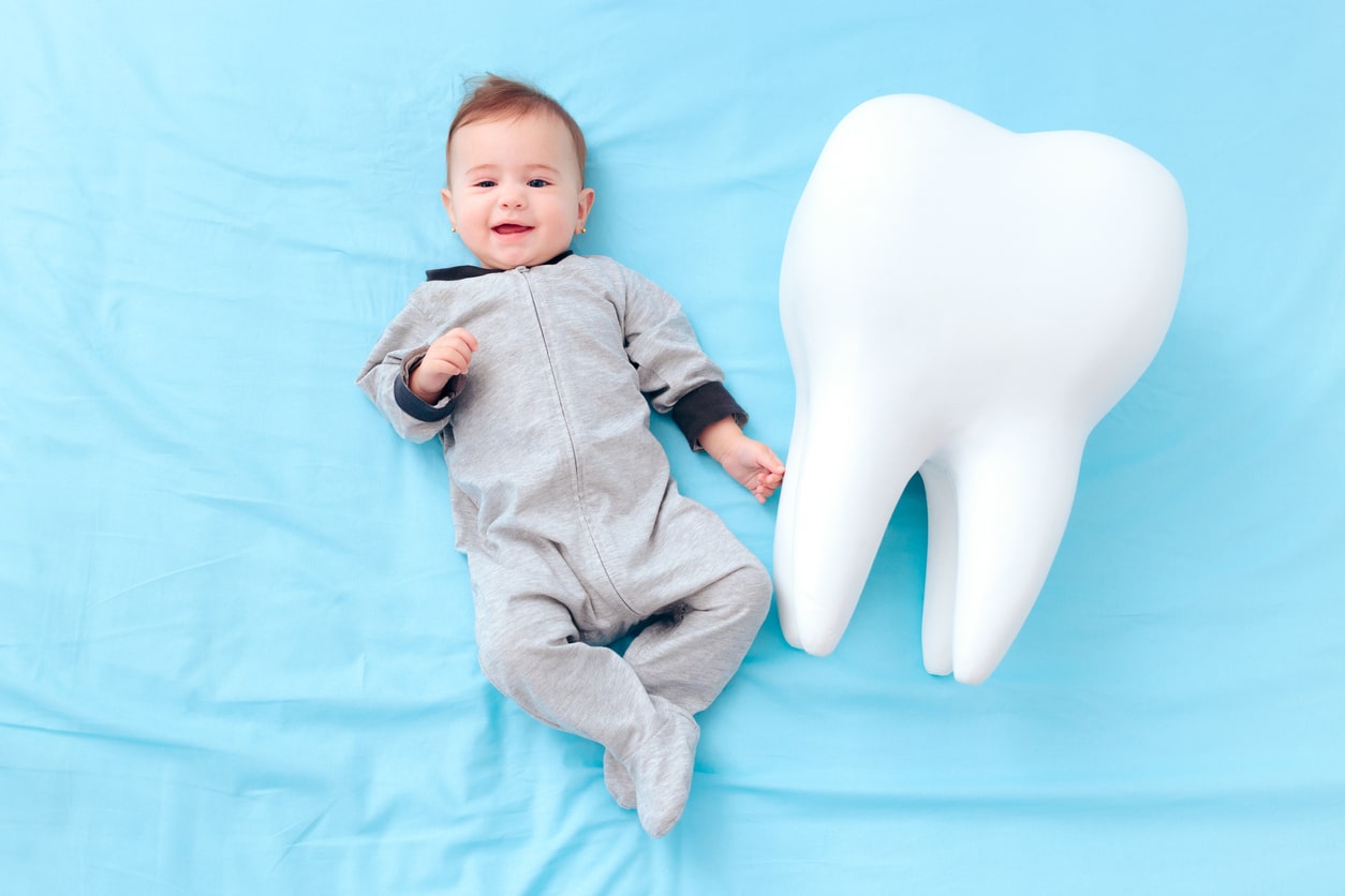 Cute baby in gray onesie lying on blue blanket next to giant white tooth