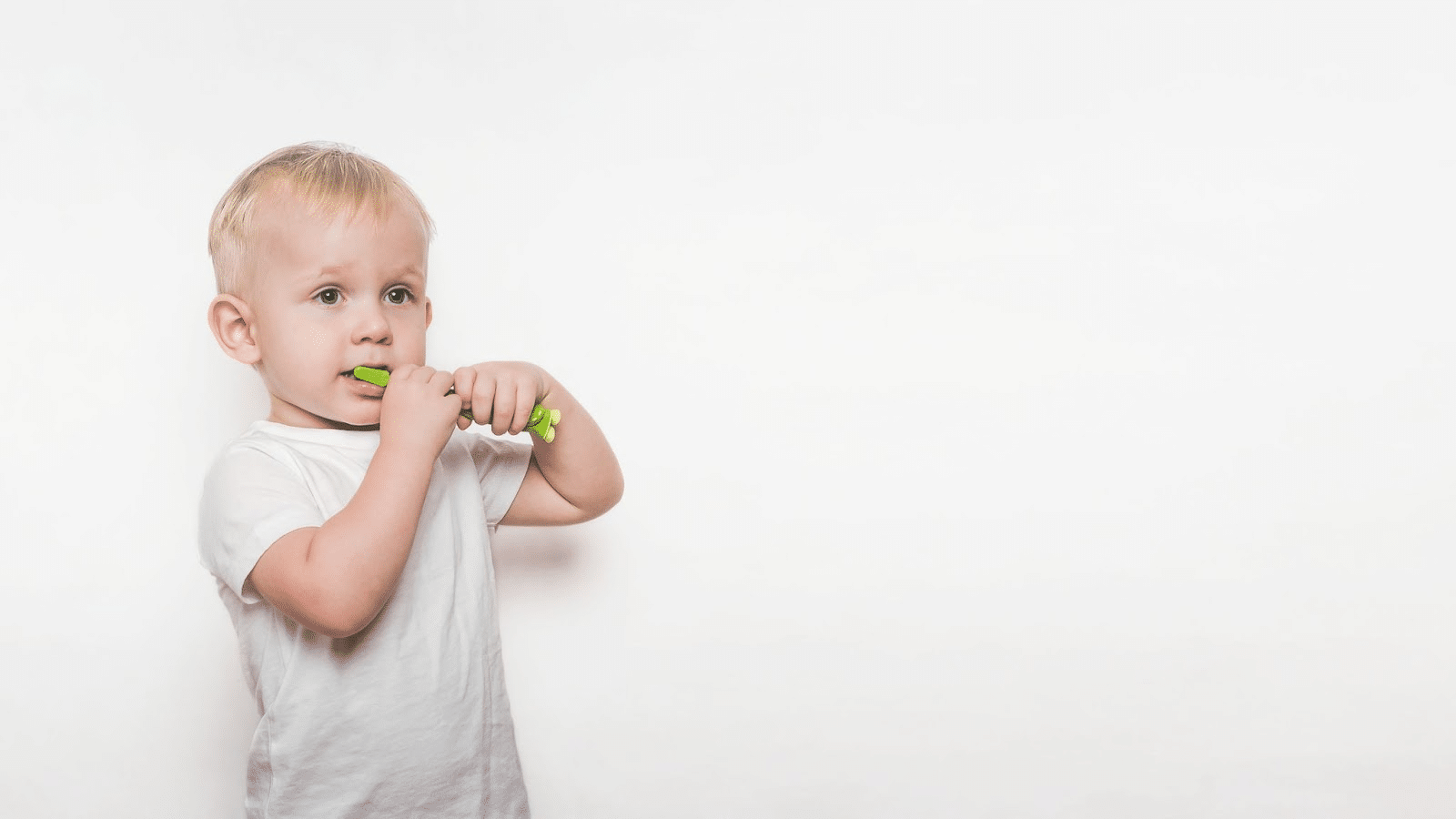 Toddler boy in white shirt brushing teeth with green toothbrush while standing in front of white wall