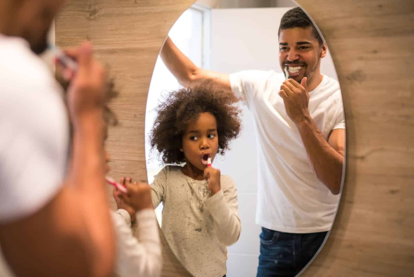 Cute little girl and handsome dad brushing teeth together in front of oval mirror