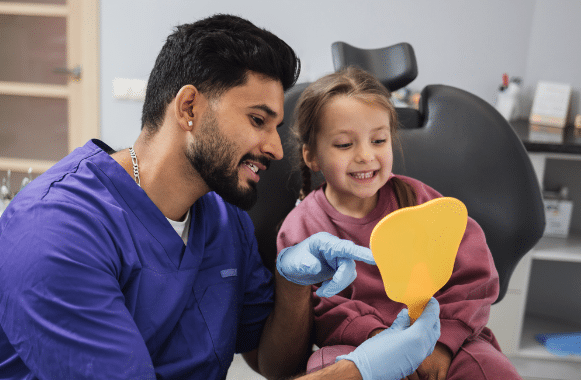 Cute little girl looks at her smile as dental assistant holds yellow mirror for her