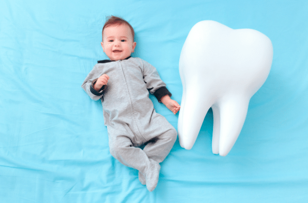 Smiling baby in gray onesie lies next to giant toy tooth on blue blanket